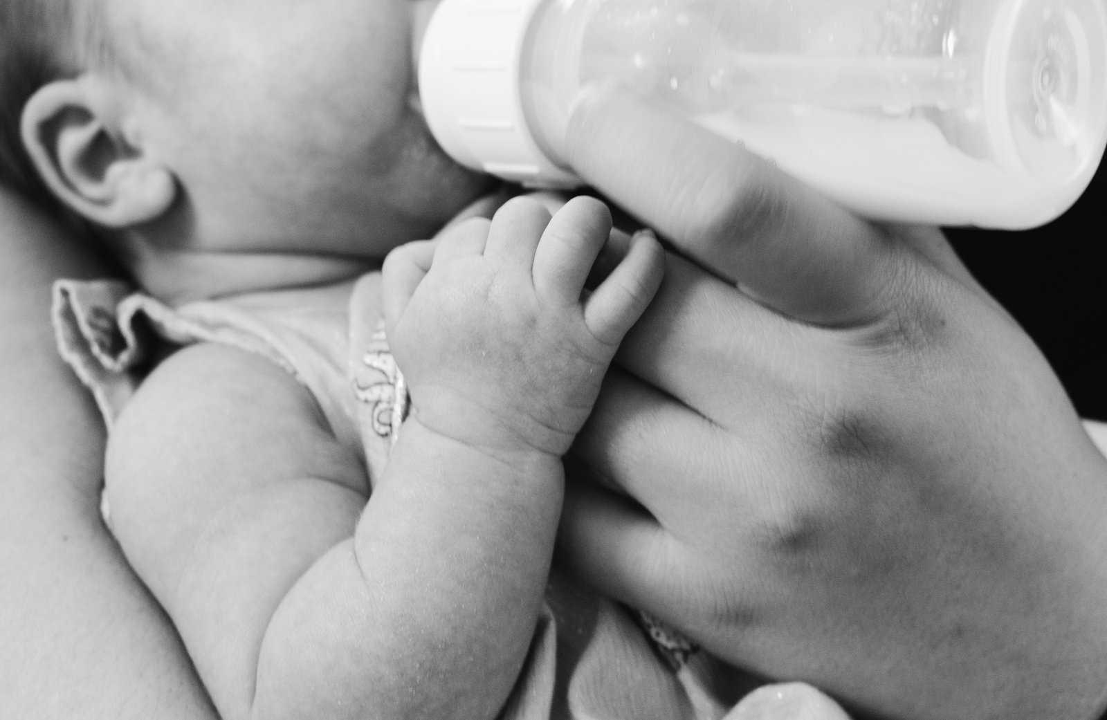 A newborn feeding from a bottle, hand wrapped around the parent's finger